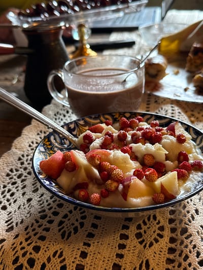 A close-up shot shows a bowl of fruit salad topped with wild strawberries and possibly pears or apples, garnished with white powder, placed on a crocheted doily. Next to the bowl is a glass mug of hot chocolate with a spoon. In the background, a cezve sits near a glass cake stand filled with berries, and a slice of cake is visible on a plate. The scene is set indoors, on a wooden surface, suggesting a domestic environment. The location is not discernible from the image itself.