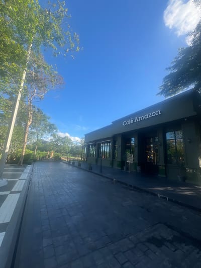 The image shows the exterior of a "Café Amazon" in Phang-nga, Thailand. The building has a modern design with large glass windows and a dark-colored facade. The café's name is prominently displayed in white lettering on a long, illuminated sign. Outside the café, there is a paved walkway with a brick pattern, and some greenery, including trees and bushes, can be seen. The sky is bright blue with a few wispy clouds, indicating it is daytime with good weather. The scene appears to be a quiet, ordinary day at the café, with no people or specific activities visible. The overall impression is one of a clean, well-maintained establishment in a natural, possibly tropical, setting.
