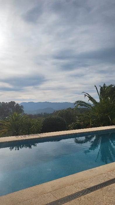 This image depicts an outdoor scene within Polígon Industrial De Calvià, Spain. The foreground features a rectangular swimming pool with visible blue mosaic tiling and clear water, which reflects parts of the sky and foliage. The pool is bordered by light-colored, textured coping stones. A dark, linear grated drain runs parallel to the pool's edge on a light-colored tiled deck in the lower right quadrant. Beyond the pool, a landscape of dense green vegetation, including multiple palm trees and various bushes, extends across rolling terrain. In the background, a range of forested mountains rises against a partly overcast sky. The sky exhibits white and grey cloud formations, with a prominent bright light source located in the upper left quadrant. No individuals or specific actions are discernible in the frame.