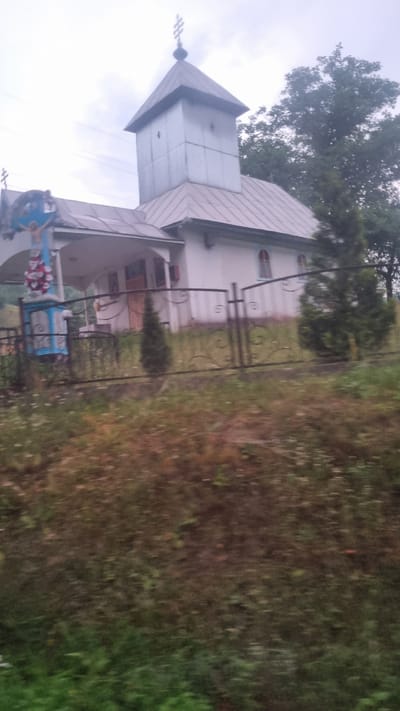The image shows a white church with a metal steeple in Kostrynska Roztoka, Ukraine. A cross is visible on top of the steeple and another smaller cross is positioned on the left side of the church. In front of the church, there is a blue and white crucifix with a statue of Jesus Christ. A black metal fence encloses the church property. There are two small evergreen trees in front of the church and a larger tree on the right side. The ground is covered with grass and some wildflowers. The sky is overcast. The image appears to have been taken from a moving vehicle, as evidenced by the motion blur in the foreground.