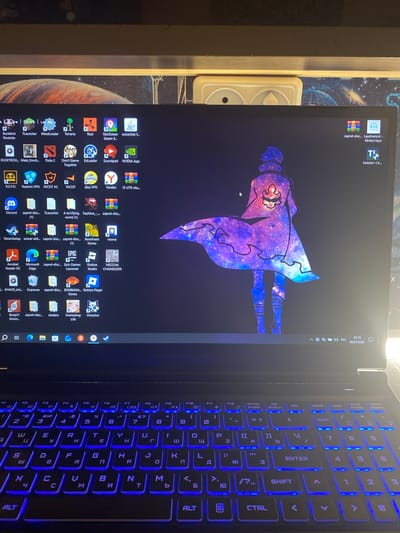 The image shows a close-up view of a laptop with a dark background on the screen. The screen displays a desktop with numerous application icons and a wallpaper featuring a stylized character dressed in a purple and blue galaxy-themed outfit. The character is depicted in a dynamic pose, seemingly floating or flying.

On the right side of the laptop, the keyboard is illuminated with vibrant blue backlighting. The keys are arranged in a standard QWERTY layout, with some Cyrillic characters also visible, indicating the potential for a Russian language setting. The keyboard is the primary focus of the right half of the image, showcasing the glowing keys.

The bottom right corner of the screen displays system information, including the time "22:14" and the date "09.07.2026". The presence of "PYC" suggests the Russian language is in use.

The scene is indoors, likely a room in a home or office, given the presence of the laptop and the ambient lighting. The overall mood appears casual, perhaps indicating the user is engaged in gaming or other computer-related activities. The distinct wallpaper suggests a personal preference for fantasy or anime aesthetics. There are no visible people in the image.