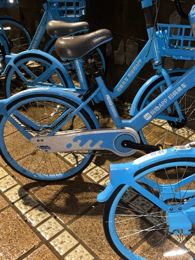The image depicts multiple blue share bicycles, identified by "HELLO DESIGN" and "哈啰出行" (Hello Bike) branding, parked on wet, tiled pavement in Yanqiao, China. The bicycles are bright blue with black seats, baskets, fenders, and chain guards featuring a white and light blue wave pattern. Water droplets are visible across all surfaces of the bicycles, including the seats, frames, and baskets, indicating recent rain. The paved ground, composed of light brown or beige square tiles with darker grout lines, is also wet and reflects ambient light, creating shadows from the bicycle components. The background is dark and indistinct, suggesting an urban environment at night or in low-light conditions. No individuals are present in the scene.