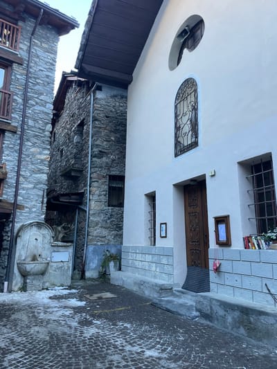 The image captures a narrow, traditional street or alleyway in Courmayeur, Italy, characterized by rustic stone architecture and a contrasting lighter facade. The scene is quiet and appears to be daytime, with clear lighting.

On the left side, a multi-story building features a light-colored upper section with distinct architectural elements, transitioning to dark, rough-hewn stone masonry at its base. The upper part of this building includes a semi-circular cutout in the wall, possibly housing a speaker or light. Below this, an arched window is adorned with intricate black wrought-iron grilles. A dark brown wooden door with carved details serves as the main entrance, accessible via a small set of light-colored stone steps with a dark doormat. Above the door, a small rectangular window with an unreadable sign is present, and on the windowsill to its right, a stack of colorful books is visible. Below the door, another small framed sign displays what appears to be "HB" or similar text. Further down, a smaller window with simple grilles is set into the wall.

Opposite this building, on the right side of the alley, stands another structure made entirely of dark stone, which also has a small barred window. A metal pipe runs horizontally along its wall. In the foreground on the right, a traditional stone water fountain with a metal spout and a large basin is situated, adding to the historic ambiance. The ground is paved with small, dark, possibly cobblestone-like material, showing some lighter, wet patches or melted snow/ice. A small amount of greenery grows at the base of the lighter building. No people are visible, and no specific event is taking place, presenting a tranquil streetscape.