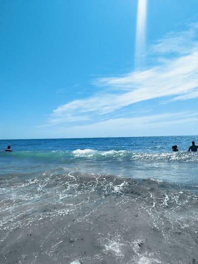 A bright, sunny day at the beach in Rota, Spain. The image captures a wide view of the ocean and sky. In the foreground, the shallow water laps against a sandy, dark gray seabed, with white foam from the waves creating patterns on the surface. A medium-sized wave breaks in the middle ground, its crest frothy and white. The sea is a deep blue, stretching out to the horizon. In the distance, several figures are visible in the water. On the left, a single person is swimming, their head and shoulders above the surface. Towards the right, two more figures are present in the water. Further out in the sea, a few thin, vertical structures, possibly wind turbines or other markers, are visible on the horizon line. The sky is a vibrant, clear blue, with scattered white clouds, some wispy and thin, others more substantial. A bright, almost white, vertical beam of light cuts through the sky from the upper right quadrant of the image, suggesting either a sunbeam or a lens flare. The overall impression is one of a pleasant day at the seaside.