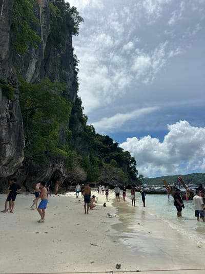 The image shows a group of people on a sandy beach with a rocky cliff face covered in lush green vegetation on the left. The sky above is a mix of blue and white clouds. In the background, several longtail boats are moored in the clear, shallow water.

The people on the beach appear to be engaged in some form of activity, possibly a group exercise or a performance, as they are positioned in a line facing the water, some with their arms outstretched. Several individuals are wearing swimwear.

The scene suggests a tropical destination, with the characteristic longtail boats and rocky karsts common in places like Ton Sai, Thailand. The lighting indicates it is daytime, likely mid-morning or afternoon, with the sun illuminating the beach and water. The weather appears pleasant with a mix of sun and clouds. The overall mood seems to be relaxed and recreational. There is no visible text in the image.