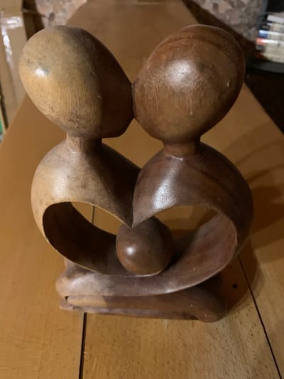 An abstract wooden sculpture depicts two figures in an embrace, their heads tilted towards each other as if sharing a kiss. The sculpture is made of light and dark wood, with the lighter wood forming the upper portions of the figures and the darker wood creating a heart-like shape in the lower section. A small, round object, possibly representing a heart or a child, is nestled between the figures within the heart shape. The sculpture rests on a wooden surface, which appears to be a table. In the background, out of focus, are shelves stacked with books and other objects, suggesting an indoor environment. The lighting is dim and warm, casting shadows on the wooden surface. The sculpture is situated on a table in what appears to be a room in Mepiskalaki, Georgia.