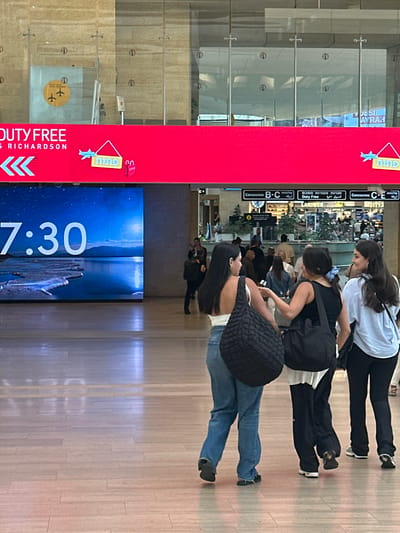 The image captures a scene in Terminal 1 of Ben Gurion Airport, located in Tzafria, Israel. A large, bright red digital display board dominates the upper portion of the frame, featuring the text "DUTY FREE" and a shopping bag icon. Below this, a digital clock displays "7:30" against a serene blue background of water and mountains. In the foreground, three young women are walking away from the camera. The woman on the left wears a white top and blue jeans, carrying a large black quilted bag. The middle woman has dark hair and wears a black top and dark pants, also with a black bag. The woman on the right is in a light blue shirt and black pants, with a black bag slung over her shoulder. They are walking on a polished wooden floor in what appears to be a modern airport terminal. In the background, airport signage indicates concourses and a "Duty Free" shop. Other travelers are visible in the distance, adding to the bustling airport atmosphere.