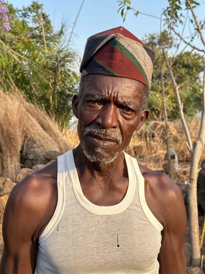 An elderly Black man gazes intently, wearing a multi-striped hat and a white tank top with small holes. The background includes trees, foliage, and a weathered, fibrous structure. He has a graying beard and wrinkles, evidence of age. The lighting suggests an outdoor setting, possibly during the day, with natural sunlight casting shadows and highlighting textures.