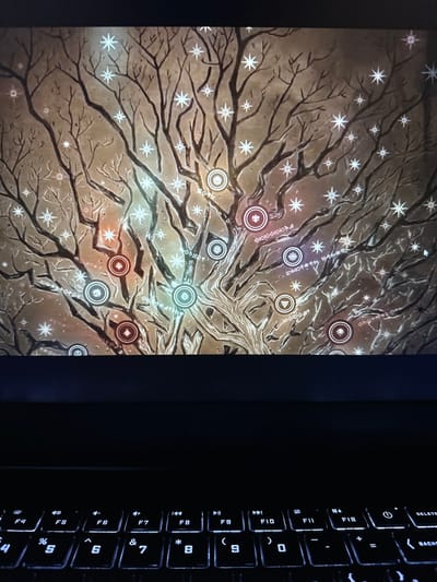 The image shows a close-up, slightly angled view of a laptop screen displaying a stylized, illustrated tree with many branches. The tree has a sepia-toned background and is adorned with white, star-like symbols and glowing circular icons. The branches are dark and intricate, resembling a winter scene. The lower part of the image shows a backlit keyboard of a laptop. The screen content is the primary focus, with the keyboard serving as a framing element. There are no people visible in the image, and no identifiable landmarks or clues about a specific location. The image does not provide any information that would allow for the determination of its location in Vancouver, United States of America, or any other city.