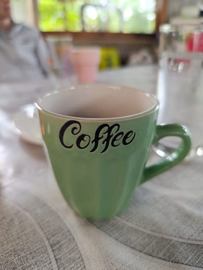 A light green ceramic mug with the word "coffee" in black script is positioned in the foreground of the image. The mug is filled with a dark liquid, likely coffee. Behind the mug, a white saucer is partially visible. In the background, out of focus, are several objects including a clear glass with liquid, a pink cup, and a blurred figure of a person wearing a gray jacket. The scene appears to be set indoors, possibly in a cafe or home, with a patterned tablecloth beneath the mug. The lighting suggests it is daytime. The image does not contain information related to Mladinovo, Bulgaria.