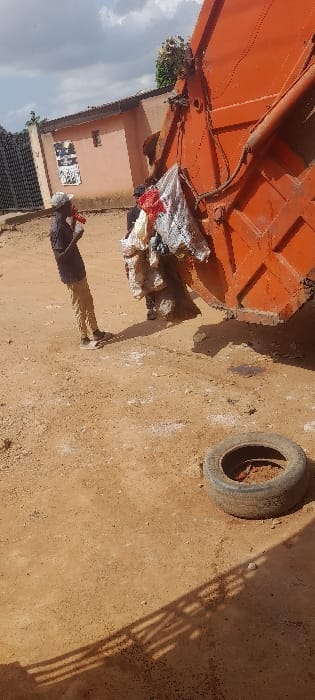 The image displays an outdoor waste collection scene in Ashipa, Nigeria, under daylight with strong shadows indicating sunlight. An orange rear-loader garbage truck occupies the right side of the frame, with its compaction mechanism actively loaded with multiple white, red, and miscellaneous bags of refuse. On the left, a male individual stands facing the truck, dressed in a dark short-sleeved shirt, light brown trousers, and a white baseball cap, holding a red object near his mouth. The lower legs of a second person are visible directly beneath the rear of the garbage truck. The ground consists of compacted, light brown earth with scattered dark particles. In the lower right foreground, a single weathered black rubber tire rests flat on the ground, partially filled with dirt. The background includes a light brown residential-style building with a dark roofline, featuring posters affixed to its wall, and a black metal gate or fence to its left. Distinct linear shadows, possibly from a fence, stretch across the lower left portion of the ground.