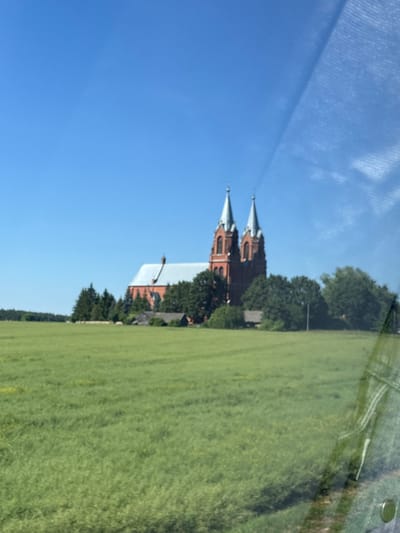 The image displays a rural landscape under a clear blue sky. In the mid-ground, a large, red brick church with twin spires stands prominently, surrounded by lush green trees. In the foreground, a vast field of green crops stretches across the scene. The image appears to have been taken from a moving vehicle, as evidenced by a slightly blurry, reflective surface that overlays a portion of the sky and the right side of the frame. The overall impression is one of a peaceful, pastoral setting in the Belarusian countryside, specifically within the Васілішкаўскі сельскі Савет region.