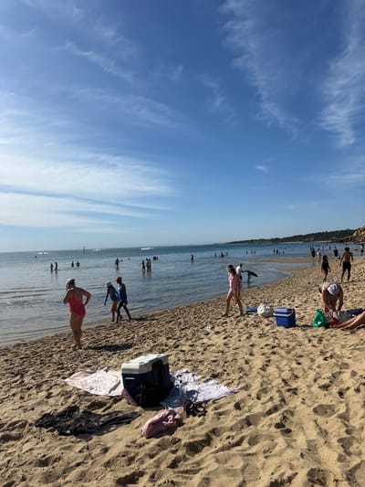 A sunny beach scene shows people enjoying the ocean and sand. Some individuals are swimming in the water, while others relax on the beach. Beach essentials like coolers and bags are visible. The sky is bright blue with wispy clouds. The sand is a light tan color. The overall atmosphere is relaxed and summery.
