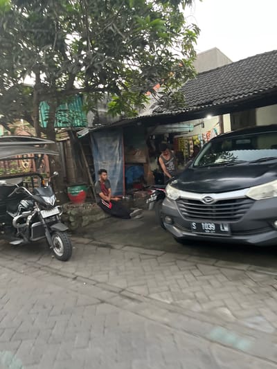 The image shows an outdoor scene in Karangtanjung, Indonesia. In the foreground, a motorcycle is parked on a brick-paved surface. Behind the motorcycle, a dark-colored car is parked in front of a small shop or stall. Two men are visible in the scene. One man, wearing a dark t-shirt, is leaning towards the car, possibly working on it or inspecting it. He appears to be engaged in some activity related to the vehicle. The second man is sitting on the ground to the right of the shop, with his legs crossed and his arms resting on his knees. He is looking towards the car and the other man, and his expression suggests he is observing or waiting.
The shop appears to be stocked with various items, suggesting it might be a small convenience store or workshop. There are shelves with products visible inside. A large, red and green drum is also present near the seated man. A tree with green leaves partially obscures the view of the shop and the surrounding buildings. The overall setting suggests a casual, possibly commercial, environment.
The lighting in the image is somewhat diffused, indicating it might be overcast or the time of day is either early morning or late afternoon. There is no strong sunlight. The brick pavement suggests an urban or semi-urban setting.
Visible text includes a license plate on the car which reads "S 1039 LM". There is also some text on the red and green drum, though it is partially obscured and difficult to read clearly. The second license plate on the motorcycle is also visible and reads "P 2348 B...".