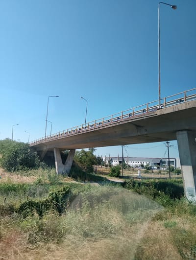 The image captures a concrete overpass spanning a dry, grassy area. The overpass has yellow railings and is supported by two large concrete pillars. Several streetlights are visible along the overpass and in the surrounding area. In the background, there are industrial buildings with white facades and a clear blue sky. The scene appears to be in a suburban or industrial outskirts area of the Municipal Unit of Echedoros, Greece, likely on a sunny day. There are no people or vehicles immediately visible in the foreground or on the overpass.