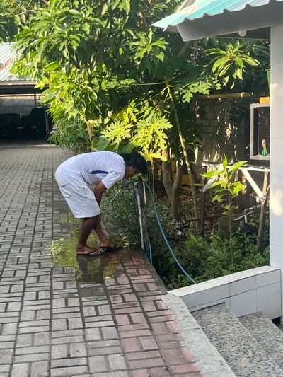 A person wearing a white t-shirt and white shorts is bending down to drink water from a hose in front of a building. The person is standing on a paved walkway that is wet from the hose. Green trees and plants are visible in the background. The scene appears to be outdoors in a tropical or subtropical climate, possibly Pemenang Timur, Indonesia, given the vegetation and building style. The person's posture suggests they are either taking a drink or washing their face.