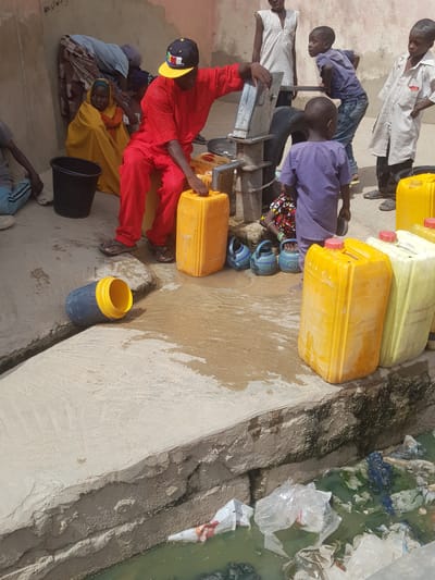 In Dutsinma A, Nigeria, a group of people, including children, are gathered around a hand pump filling yellow jerrycans with water. The environment shows signs of poor sanitation, with debris and murky water visible in an adjacent ditch. People are sitting and standing, with filled jerrycans nearby. Some are wearing traditional clothing, while others are in more casual attire. A black bucket sits on the ground.