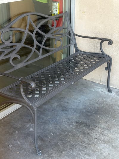 A dark metal bench with ornate scrollwork on the backrest and a lattice-patterned seat and backrest is positioned outdoors on a concrete surface. The bench is facing left, with its right armrest visible against a textured, light-colored wall. To the left of the bench, a large glass window is present, reflecting the interior of a building, which includes a glimpse of a fire extinguisher and some boxes. The concrete ground appears worn and slightly stained. The scene is static, with no individuals or discernible activity. The lighting suggests it is daytime, with shadows cast by the bench.