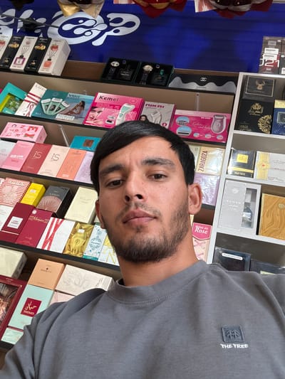 A close-up selfie shows a young man with dark hair and a beard in a gray sweatshirt. He is positioned in front of shelves stocked with various cosmetic products, including perfumes, lotions, and hair care items. The shelves are organized and brightly lit. The background reveals a blue wall with some decorative elements. The man appears to be in a retail setting, possibly a beauty store or a section of a larger shop, located in Yovon, Tajikistan.