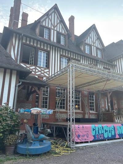 The image depicts a half-timbered building, possibly a historical residence, in Gueutteville, France. In front of the building, a temporary stage has been set up with a light-colored canopy supported by silver metal scaffolding. A pink banner with large teal lettering that reads "CIRCUS & CO" is draped across the front of the stage. Various logos are visible on the banner, suggesting sponsorships. To the left of the stage, a small, colorful carousel or merry-go-round with sea creature figures and painted scenes is present, along with a potted plant. Yellow electrical cords are coiled on the ground near the stage. The building's architecture features prominent exposed wooden beams against white plaster, with red brick sections visible. Several windows, some open, are apparent on the facade. The sky is overcast.