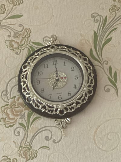 A decorative analog wall clock with a silver ornate frame is centered on a textured wallpaper. The wallpaper has a floral pattern in muted green and beige tones. The clock face is white with black Arabic numerals and hands. The hour hand points to approximately 10, and the minute hand points to approximately 5. The second hand is red and points to just past 6. The clock has a dark brown bezel. The image is a medium shot of the clock and its surroundings, taken from a slightly downward angle. There are no people or actions depicted in the image. The scene appears to be indoors, likely in a residential or public space.