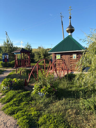 The image depicts a small wooden chapel with a green roof and a golden dome topped with a cross. Adjacent to the chapel is a small red arched bridge leading over a dry well. To the left, there is a red wooden structure with a roof, possibly a kiosk or shelter, with a bell inside. In the background, trees and a power pole are visible under a clear blue sky. The scene appears to be in a rural area, likely Pobedinka, Russia, given the presence of Orthodox Christian symbols like the chapel and crosses. There are no people or animals visible in the image. The environment is overgrown with grass and bushes, suggesting a natural or semi-natural setting.