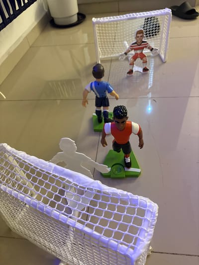 The image depicts a tabletop foosball setup on a light-colored, reflective surface, likely a table or floor. Two foosball goals, constructed from white mesh netting and plastic frames, are positioned on opposite ends. Four foosball players are visible: one wearing a red and white goalie uniform within the left goal, one wearing a blue jersey and black shorts, and another wearing an orange jersey and black shorts facing the right goal. A flat white soccer player figure is positioned near the right goal. The foosball players are connected to green bases with knobs. Additional details include a black slide, and a black and white striped object on the left side of the image. No geographic information is discernible from the image.