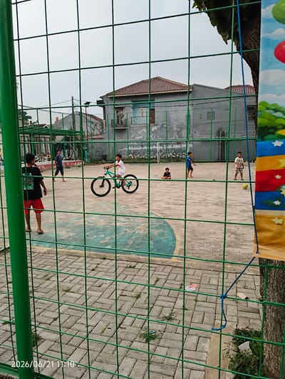 A group of young boys are playing soccer on an outdoor concrete court in Bojong Gede, Indonesia. One boy rides a bicycle in the foreground. The court is surrounded by a green chain-link fence, with houses and trees visible in the background. The sky is overcast.