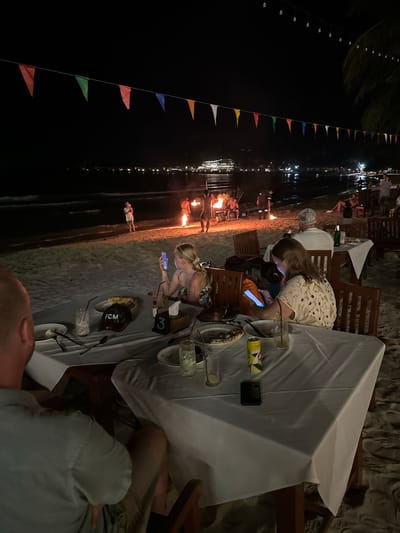 The photograph captures an outdoor evening scene, likely a beachfront dining area, in Hat Khai Muk, Thailand. A large table, covered with a white tablecloth, dominates the foreground. Several people are seated around the table. One person is bending over, focused on their phone. Another person is also on their phone. The table contains plates, glasses, beverages in a can, and other objects. 

In the background, a beach is visible with a line of people dancing with fire and a decorative string of colorful flags. Lights twinkle in the distance along the shoreline. The overall ambiance suggests a resort or entertainment venue on the beach at night.