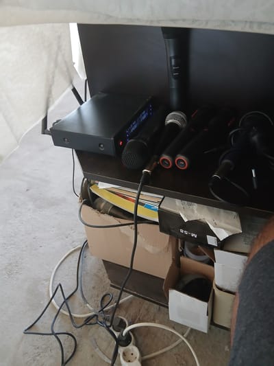 A top-down, slightly rotated shot reveals a cluttered shelf in Vetren Dol, Bulgaria. Visible on the left are two black wireless microphones and a black receiver with visible interface lights. To the right of the receiver, more microphones lie among boxes and cords, including a box labeled "M-58" and some Cyrillic text. The shelf is overflowing with items, and the floor beneath is littered with electrical cords and a power strip. A portion of a person's arm, clad in gray fabric, appears in the top right corner of the frame.