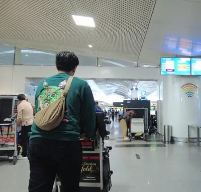 An indoor scene at what appears to be an airport, brightly lit with overhead square lights and a perforated ceiling. In the foreground, a person stands with their back to the camera, wearing a dark green long-sleeved shirt with a light green graphic on the back depicting an animated character, black pants, and a tan sling bag or fanny pack worn across the body. They are positioned behind a luggage cart, part of which is visible, featuring a poster that reads "Dreams unfold," "Sleepiestout," and "Dreamiest bed."
Further into the airport terminal, other travelers are visible. To the left, another person in a light-colored shirt is near a luggage cart. In the center-right midground, a person is seen bending over a baggage scanning machine, indicating a security checkpoint or baggage drop-off area. In the background, more people are moving through the terminal, some with luggage.
On the right side of the image, multiple digital display screens show flight information, including "FLY DUBAI" and what appears to be departure/arrival details for destinations like "BALI" and "SYDNEY." Below the screens, a white column features a colorful rainbow-arch symbol resembling a Wi-Fi or signal icon. The overall setting suggests a busy, modern airport during the daytime.