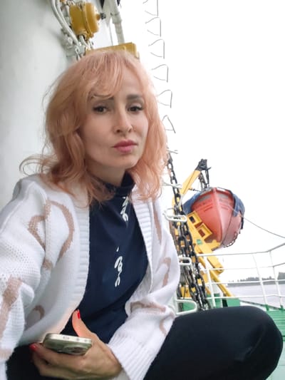 A woman with salmon-pink, wavy hair and dramatic makeup sits on a ferry deck in Nikol'skaya Gora, Russia. She is wearing a white cardigan with a tan pattern and a dark blue turtleneck. Her left hand, with red nail polish, holds a gold-colored smartphone. In the background, a red lifeboat is suspended from a yellow davit, with a thick chain hanging beside it. The railing of the ferry is visible, and beyond that, water and a distant shoreline with trees can be seen under an overcast sky.