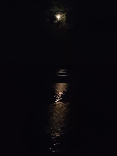 A nighttime scene shows the moon, partially obscured by clouds, reflecting on the water. The location is Krasnoe (Uleiminskoe S/P), Russia, based on the provided location details. The image focuses on the light of the moon creating a bright, shimmering path across the water surface. The rest of the scene is very dark. There are no people, subjects, or interactions immediately visible in the image.