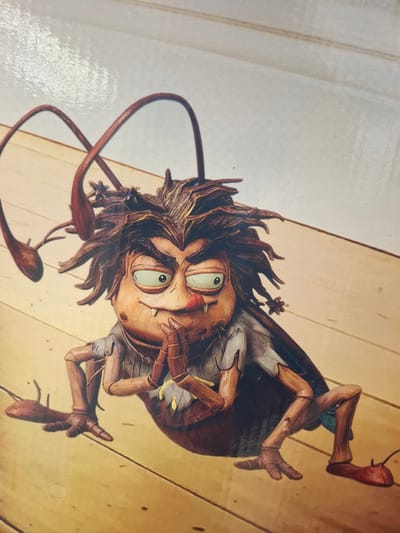 This is a close-up, low-angle shot of a stylized, anthropomorphic insect character. The character appears to be a puppet or figurine with a human-like face and insectoid body parts. Its skin is a mottled brown and tan, with large, green, wide-set eyes that give it a surprised or mischievous expression. The character has spiky, dark brown hair and prominent, segmented antennae that curve downwards. It is wearing a light-colored shirt with its sleeves rolled up and what appear to be dark brown, segmented legs and feet, ending in what resemble small, pointed shoes.
The insect character is positioned on a surface that looks like light-colored wooden planks, possibly a floor or tabletop. The background is a blurred, warm-toned surface, also appearing to be wood. The lighting is soft and even, suggesting an indoor setting, possibly in daylight. The overall impression is whimsical and a bit unsettling due to the character's unusual design and expression. There is no visible text in the image. The context of Beylikdüzü, Turkey does not provide any specific visual cues within the image itself.