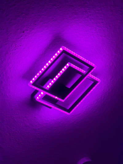 This is a close-up, overhead, indoor shot of a modern, square-shaped light fixture mounted on a textured, dark purple wall. The fixture is composed of two overlapping squares, each outlined with a strip of bright pink LED lights. The lights cast a strong purple glow on the surrounding wall, creating shadows that emphasize the fixture's geometric design. The wall's textured surface is clearly visible, with small bumps and imperfections adding depth. No people or other objects are clearly discernible in the frame. The lighting suggests this is a dimly lit room, likely a home or commercial interior. The specific location in Dorsten, Germany, is not visually represented in the image.