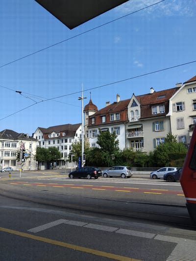 The image captures a street scene in Solothurn, Switzerland, likely near a tram stop. In the foreground, asphalt pavement with yellow and red lane markings is visible, along with tram tracks. Several cars are present on the road: a black sedan, a silver hatchback, and a dark gray hatchback. A portion of a red tram or train is on the right side of the frame.
In the mid-ground, a multi-story building with a red-tiled roof and cream-colored facade stands prominently, featuring multiple windows. Adjacent to it, another building with a similar architectural style is visible, as is a taller building on the left with a white facade and numerous windows. Trees and dense green foliage line the sidewalk in front of the buildings.
A traffic light signal and street signs are positioned at an intersection. Overhead, power lines and a suspended dark rectangular object are visible against a clear blue sky with a few wispy clouds. The overall impression is of a well-maintained urban environment on a bright, sunny day. No people are clearly discernible in the scene.