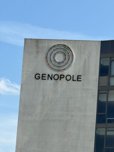 The image shows the side of a building with the name "GENOPOLE" written in large, black, sans-serif capital letters. Above the text is a circular mosaic design composed of concentric rings. Each ring is made up of small, colorful rectangular tiles in various shades of red, yellow, blue, and white. The building itself appears to be made of concrete and has a portion of a modern glass-paneled section on the right side. The sky in the background is blue with some wispy white clouds. This building is located in the city of Évry-Courcouronnes, France, and is part of the Genopole campus, a major French center for biogenetics and life sciences.