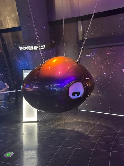An indoor exhibition space, located in Guangming District, China, features a large, dark, ovoid object suspended centrally in the frame. The object, approximately 1.5 meters in diameter, displays a metallic gradient shifting from bright orange and red at its apex to deep purple and black on its lower half. It is suspended by multiple thin cables from an overhead structure. A stylized white "eye" with a dark, horizontal, rectangular pupil-like feature is visible on the object's front surface. A small white rectangular label with the number "284" is attached to its right side.

The environment consists of dark walls adorned with a starry night sky or galaxy motif. Above the suspended object, a curved black sign with illuminated white Chinese characters reads "变化的规律是什么?" (What is the law of change?). To the left and rear of the main object, a narrow, vertically oriented, illuminated white panel stands against the dark wall. The floor is dark, glossy, and tiled with a visible grid pattern, reflecting ambient light. A circular white decal containing a green downward-pointing arrow is present on the floor in the lower-left corner. Faint reflections and partially obscured elements to the far left suggest human presence in the background.