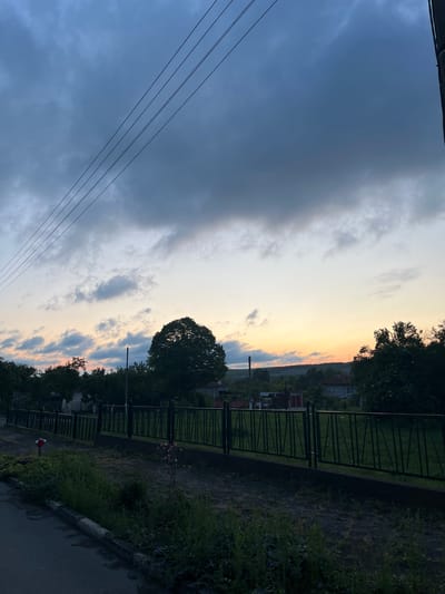 The image, likely taken in Radko Dimitrievo, Bulgaria, depicts an outdoor scene at dusk or dawn. The sky transitions from dark blue with storm clouds on the left to lighter shades with orange hues on the right horizon. Power lines stretch across the left side of the frame. A row of trees lines what appears to be a street or pathway. There's a dark metal fence running along the right side, with glimpses of buildings and vegetation beyond. A paved road and a concrete curb are visible in the lower right corner.