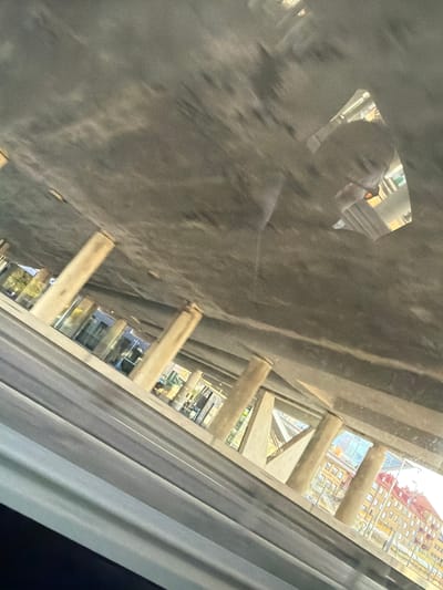 The image shows a view from under a concrete structure, possibly a bridge or overpass, looking out towards buildings and a road. Several large concrete pillars support the overhead structure. Through the gaps between these pillars, a street with cars can be seen, along with buildings featuring red and yellow facades. A reflection of a person's face is visible on the underside of the concrete structure. The scene appears to be a busy urban or suburban area.
