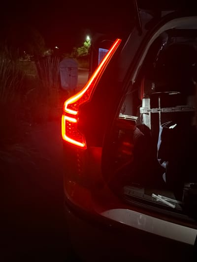 The image depicts the rear of a car with its trunk open, illuminated by the car's red taillights and some ambient light from the surroundings. The scene appears to be outdoors, possibly a parking lot or roadside, with some vegetation and a light post visible in the background. The trunk contains various objects, including what appear to be bags and possibly some equipment. The overall lighting suggests it is nighttime or dusk. The image does not contain any identifiable landmarks or clues that would place it specifically in Smyadovo, Bulgaria.