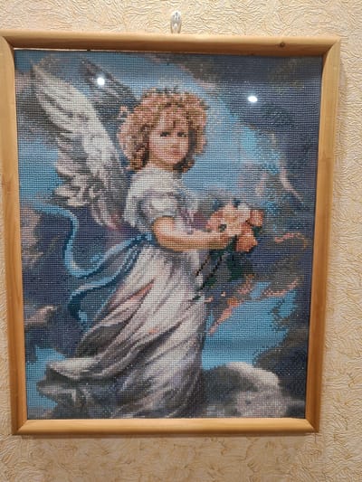 This is a framed diamond painting depicting an angel. The angel has light brown curly hair, is wearing a white dress with a blue sash, and is holding a bouquet of flowers. The background is a mix of blues and grays, suggesting a cloudy or sky-like environment. The painting is hung on a wall with a textured beige wallpaper. The image does not contain information about the city of Vichuga, Russia.