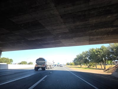The image depicts a view from under a concrete overpass on a highway in Elk Grove, California. The overpass forms a dark, textured ceiling, with visible expansion joints and some staining. Below, a bright, sunny sky is visible, with a clear blue hue.
On the highway, a large silver tanker truck is the most prominent object. The truck has "NOW HIRING SAFE DRIVERS!" written on its side in white text, along with a phone number: "816-374-7675". It also displays some hazard symbols. The truck is in the right lane, moving away from the viewer. Other vehicles are present on the highway, including a blue car and a white SUV further ahead.
To the right of the highway, a grassy embankment with scattered trees and bushes lines the road. The trees are green and full, suggesting a warm season. In the distance, some buildings are visible, and a streetlamp stands tall against the sky.
The scene suggests a daytime drive, likely during the late morning or early afternoon due to the bright sunlight and clear sky. The overall mood is one of travel and everyday highway activity. The location context is explicitly identified as Elk Grove, United States of America.