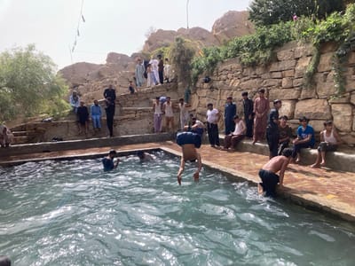 The image displays an outdoor scene in Killi Malik Sher Mohammad, Pakistan, centered on a rectangular, light blue-green pool. Approximately 25-30 males, ranging from young boys to older adults, are present. Several individuals are in the water, including one male captured mid-air, head-first, appearing to dive or somersault into the pool. Another male, shirtless, is climbing out onto the concrete edge of the pool. Other males are seated or standing along the pool's edge and on tiered stone steps and ledges integrated into a dry, rocky landscape. Many are dressed in traditional shalwar kameez, while those in or near the water wear lighter clothing or trousers. A prominent stone retaining wall, constructed from irregular light-colored stones, forms the background, behind which rocky hills with sparse vegetation ascend. A large green tree is visible on the far left. A string of incandescent light bulbs is suspended across the upper portion of the scene. The area is illuminated by bright daylight.