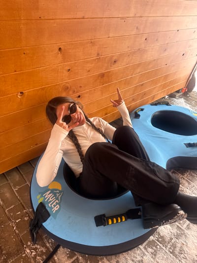 In Yeghipatrush, Armenia, a young female is depicted seated within a light blue snow tube on a partially snow-covered ground surface. She is positioned against a light brown wooden wall constructed from horizontal planks. The female has long dark hair styled in two braids, wears dark sunglasses, a white long-sleeved quarter-zip top, black trousers, and black boots. Snow residue is visible on her boots and trousers. She is smiling with an open mouth, displaying teeth. Her left hand forms a V-sign, and her right hand displays a "rock on" gesture. The snow tube features a graphic on its left side depicting mountains and a sun, with the text "MYLER" below. A black and yellow handle is affixed to the snow tube near her right foot. The ground beneath the snow consists of dark, irregular paving. A dark object, possibly another snow tube, is partially visible to the right of the primary snow tube. A dark-handled broom or brush is partially visible leaning against the wooden wall in the upper right background, adjacent to additional snow.