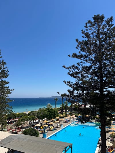 A high-angle shot captures a vibrant scene at a beachfront resort. The dominant feature is a large, bright blue swimming pool, reflecting the clear, sunny sky. Several people are visible in and around the pool, some swimming, others lounging on chairs or walking on the paved area surrounding it.
Beyond the pool, a sandy beach stretches along the turquoise waters of the ocean. Yellow umbrellas dot the beach, providing shade for beachgoers. Lush green trees and palm trees are interspersed with the beach furniture, adding to the tropical atmosphere.
In the foreground, the dark silhouette of a large tree, possibly a pine, partially obscures the view, its branches reaching across the frame. The clear blue sky dominates the upper portion of the image. The scene suggests a warm, sunny day, likely midday, with a relaxed vacation atmosphere. The location is identified as Rhodes, Greece, further reinforcing the coastal resort setting. No visible text is present in the image.