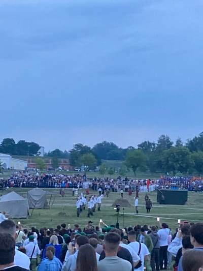 An outdoor event is taking place under a cloudy sky. A large crowd of people, many holding up their phones, are gathered in the foreground, facing a grassy field. In the middle ground, several figures in white uniforms appear to be performing or demonstrating something, while others in civilian clothing are also present. Tents and a large dark green structure are visible on the field. Further in the background, a densely packed audience is seated or standing, and beyond them, trees and some buildings are present. The overall scene suggests a historical reenactment or a public festival.