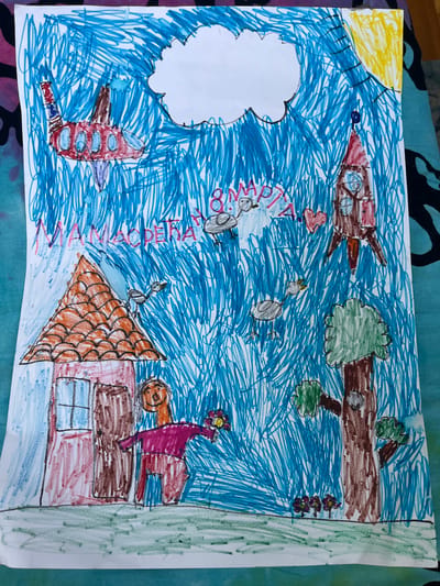 The image is a child's drawing rendered in crayon. The background is filled with scribbled blue lines representing the sky. In the upper left corner, a red and blue airplane with oval windows is depicted. To the right, a white cloud is drawn. In the upper right, a yellow sun with rays is visible. A red and brown rocket ship with windows is positioned in the upper right quadrant, pointing upwards. Below the cloud, the text "8 МАРТА" is written in pink and purple, indicating March 8th. A red heart is also present near the text. Several grey birds with red beaks are scattered throughout the sky. In the lower left, a house with an orange tiled roof and red and brown walls is drawn. A stick figure with an orange face, purple shirt, and red pants stands in front of the house, holding a yellow and red flower. The foreground depicts green scribbled lines representing grass, with small purple and yellow flowers near a brown tree with a green canopy. There is no identifiable location in Cacak, Serbia, depicted in this drawing.