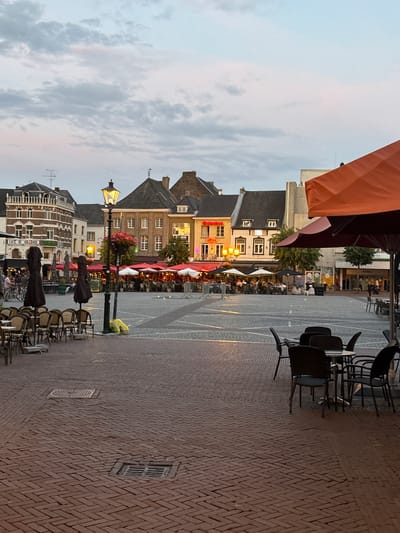 The image captures a twilight scene in Sittard, Netherlands, likely in the Markt square. The foreground is dominated by a cobblestone pavement, with several outdoor café tables and chairs set up. Some tables are empty, while others are occupied by patrons, suggesting the square is a popular gathering spot. In the mid-ground, a cluster of restaurants and cafes with outdoor seating, umbrellas, and awnings are visible. The buildings lining the square are typical of Dutch architecture, with multi-story facades and tiled roofs. A lamppost with a glowing light stands prominently in the center, casting a warm illumination on the surrounding area. The sky above is a dusky blue, with soft clouds indicating the transition from day to night. The overall atmosphere is one of a relaxed evening in a European city.