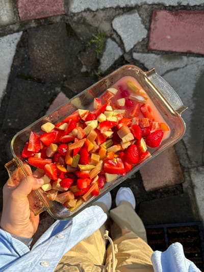 A glass baking dish, held by a person, contains a mixture of chopped strawberries, rhubarb, and possibly apples, in a light pink liquid. The dish is rectangular with vintage-style handles. The person wears light-colored pants and a light blue shirt. The background consists of a stone patio with varying textures and colors, and a small patch of grass. The photo is taken outdoors, in bright sunlight, creating strong shadows.